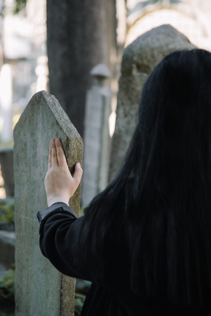 Back view of faceless female in black clothes touching old stone gravestone while spending time on cemetery on blurred background